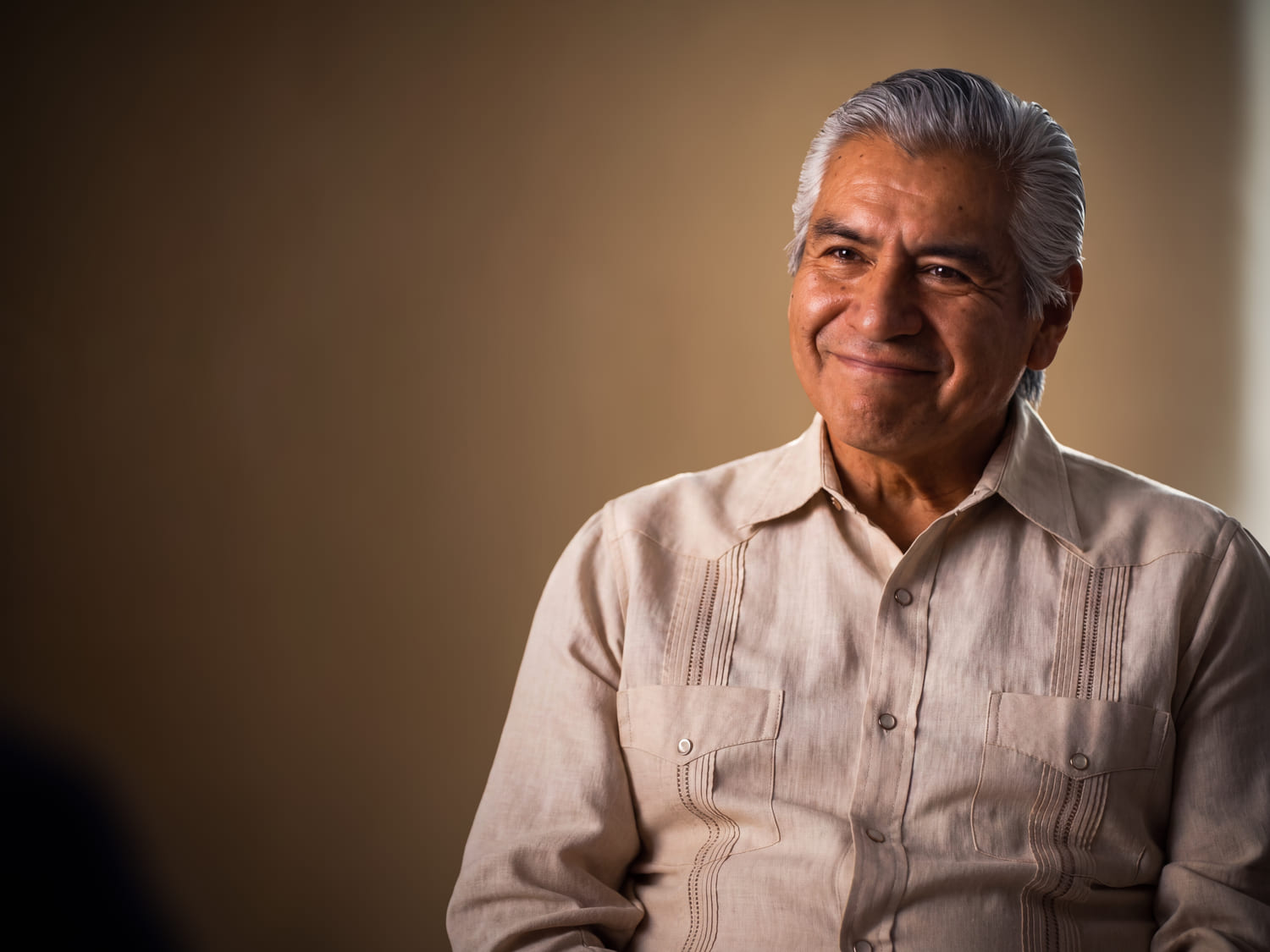 A man in his sixties sitting at ease, smiling past the camera.